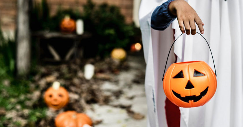 A kid dressed as a ghost trick or treating with a jack-o'-lantern candy pale